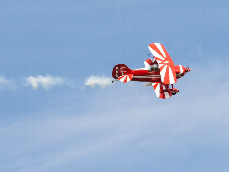 Kleines rotes Propellerflugzeug in der Luft, von der Seite betrachtet, mit weitem, blauen Himmel im Hintergrund.