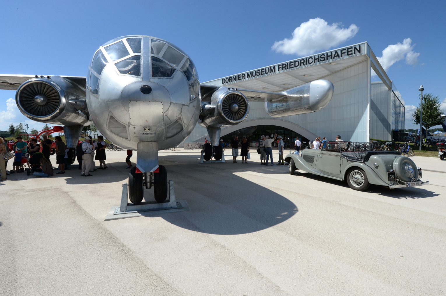 Großes silbernes Propellerflugzeug vor dem Dornier Museum Friedrichshafen, daneben Besuchergruppe und ein Oldtimer auf dem Vorplatz bei sonnigem Wetter.