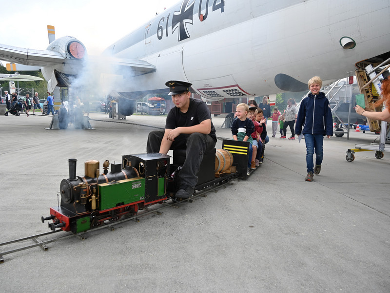 Mehrere Personen fahren mit einer Modell-Dampflock zwischen den Flugzeugen auf dem Rollfeld.