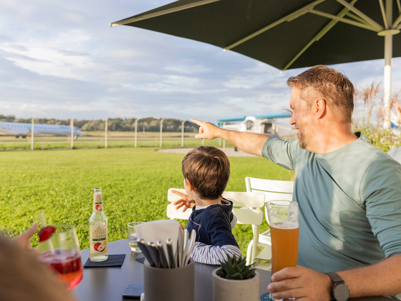 Ein Vater sitzt mit seinem Sohn im Außenbereich des Dornier-Restaurants am Tisch und zeigt auf das Rollfeld im Hintergrund.
