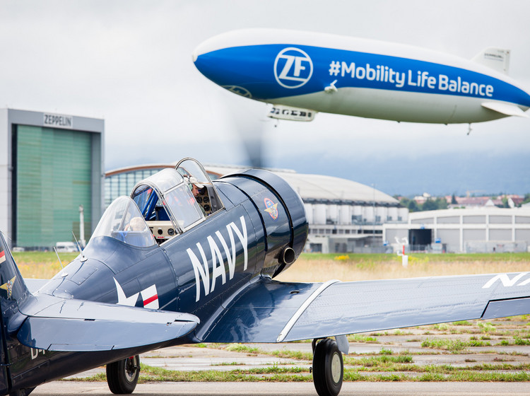 Fliegen auf zwei Arten Ein kleines Flugzeug steht mit laufendem Propeller auf der Startbahn, bereit zum Abflug. Im Hintergrund schwebt ein Zeppelin am Himmel.