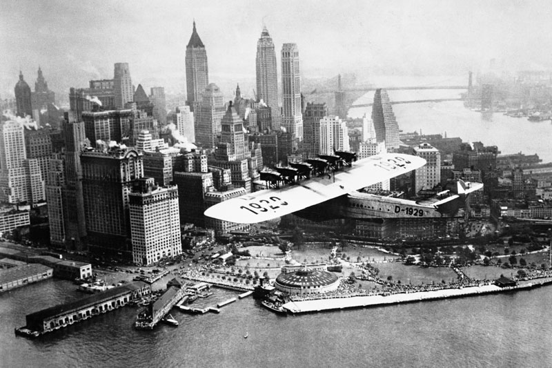 Schwarz-weiß Fotomontage eines riesigen Flugzeugs, das über die Skyline von Manhattan fliegt, im Stil einer historischen Aufnahme.